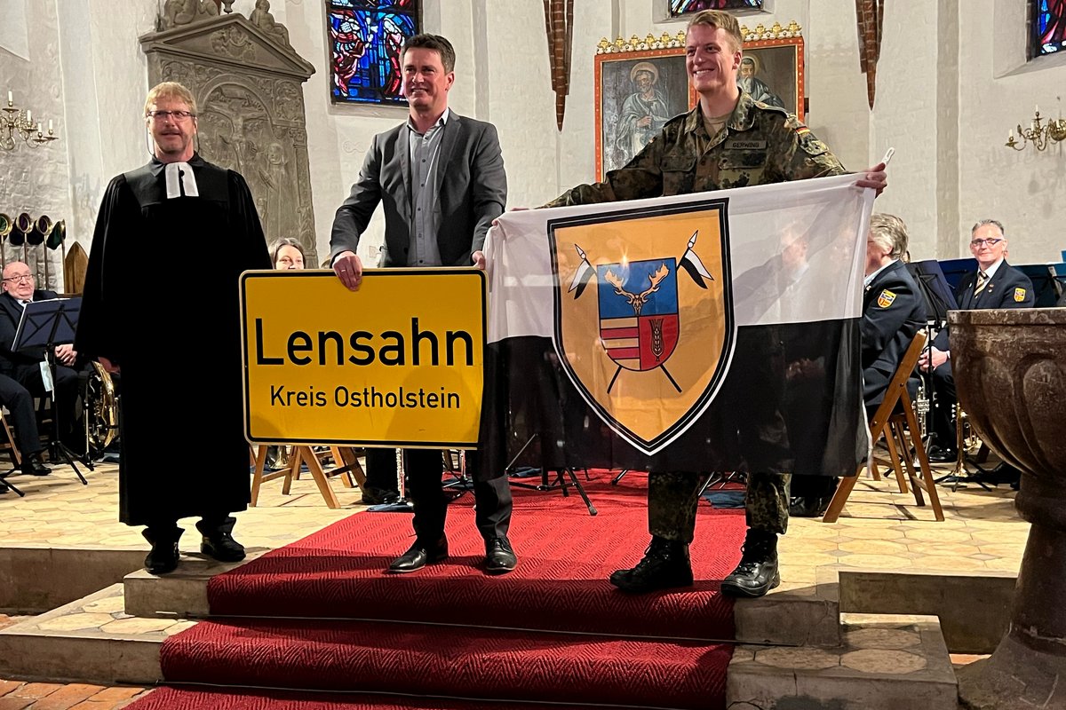 Pastor Reimann, Bürgermeister Robien und Major Gerwing stehen in der Kirch. Herr Bürgermeister Robien hält das Ortsschild der Gemeinde Lensahn in der Hand, Herr Major Gerwing hält die Fahne der Patenkompanie in der Hand.