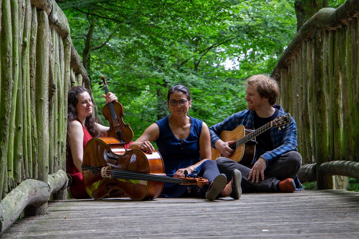 3 Personen sitzen mit Musikinstrumenten auf einer Holzbrücke im Wald